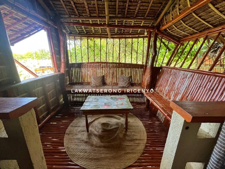 A rustic seating area is showcased, featuring a wooden table surrounded by two cushioned benches. Natural light filters through the woven walls, highlighting the textured surfaces and surrounding greenery.