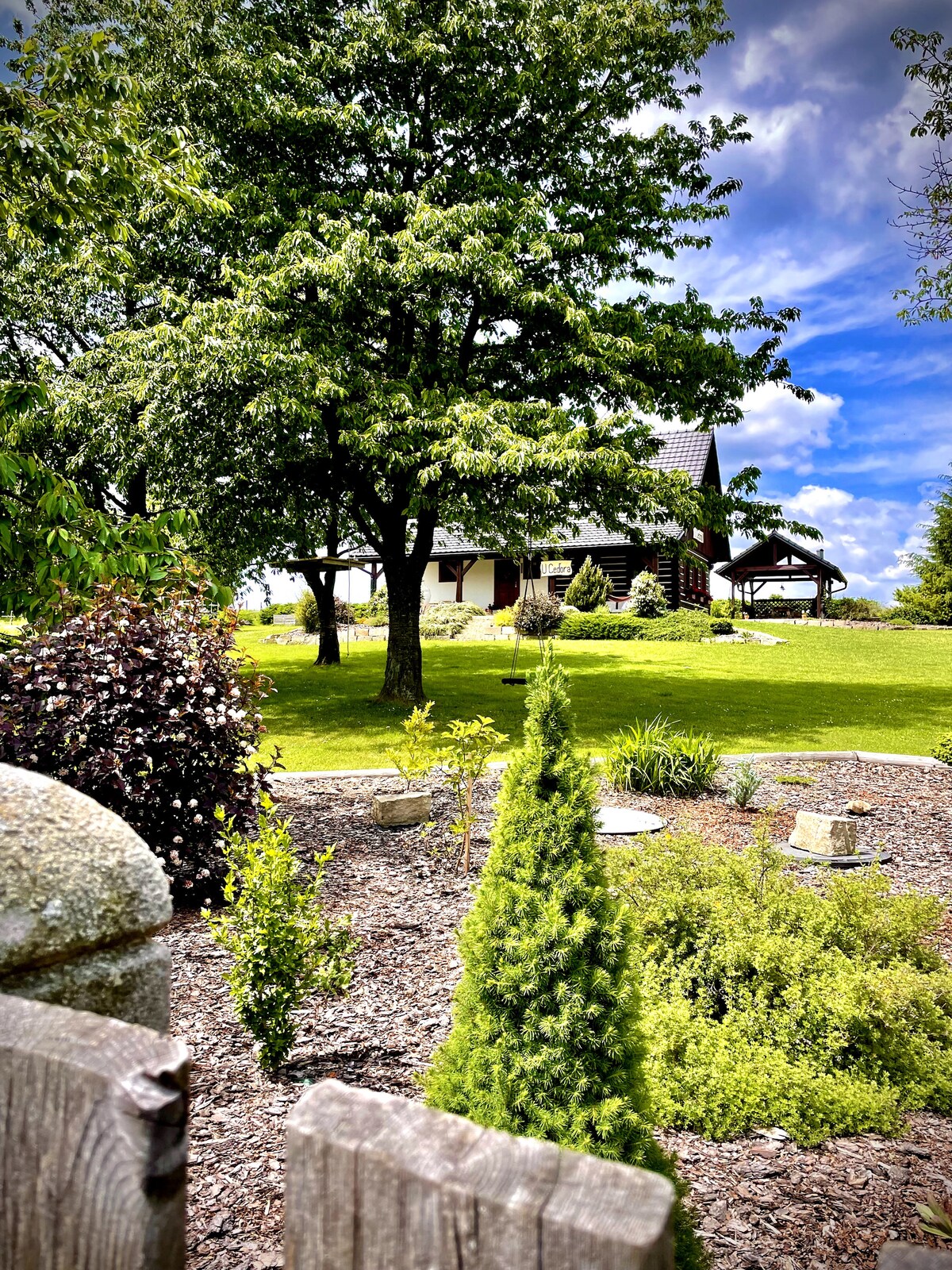 A charming wooden cabin is set against a background of lush green trees and a bright blue sky. A well-maintained garden features a mix of shrubs and small trees, creating a serene outdoor space. The scene is framed by a rustic wooden fence.