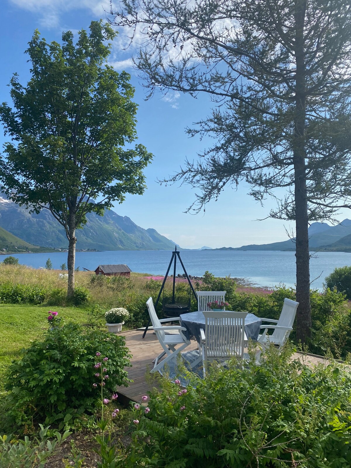 A scenic outdoor seating area is positioned on a wooden deck, surrounded by greenery and colorful flowers. White wooden chairs and a table provide a relaxing space with views of the calm water and mountains in the background, under a clear sky.