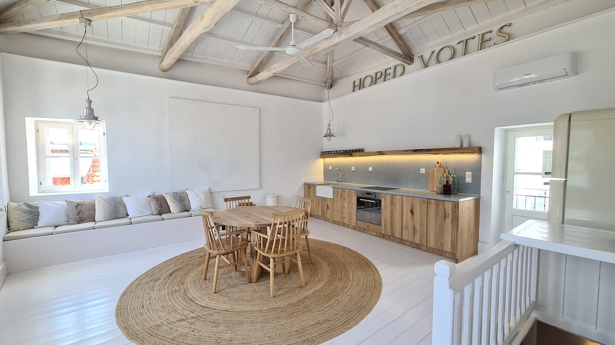 A light-filled living space features high wooden beams and a neutral color palette. A comfortable built-in seating area with soft cushions is positioned beside a round dining table surrounded by wooden chairs. A simple rug adds warmth to the polished white floor.