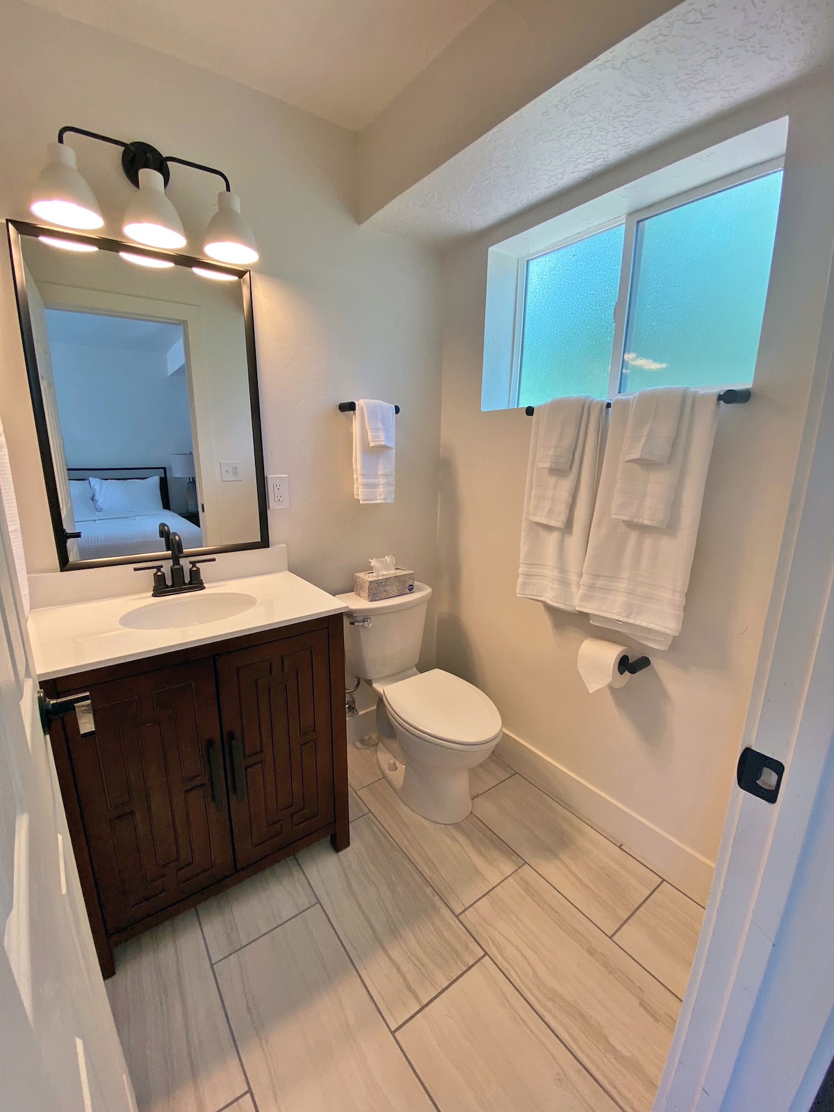 A bathroom features modern fixtures, including a dark wood vanity with a sink and mirror above. Clean towels hang neatly on towel bars. A small window allows natural light to enter, while light-colored tiles create a spacious feel.