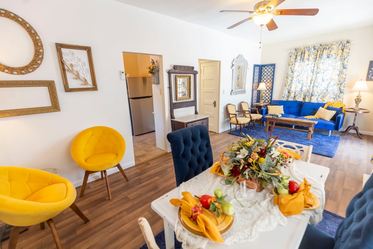 A bright and airy living space features a blend of modern and vintage elements. A dining table is elegantly set with vibrant decor, while yellow accent chairs provide a pop of color. A cozy blue sofa is positioned nearby, alongside a window adorned with cheerful curtains.