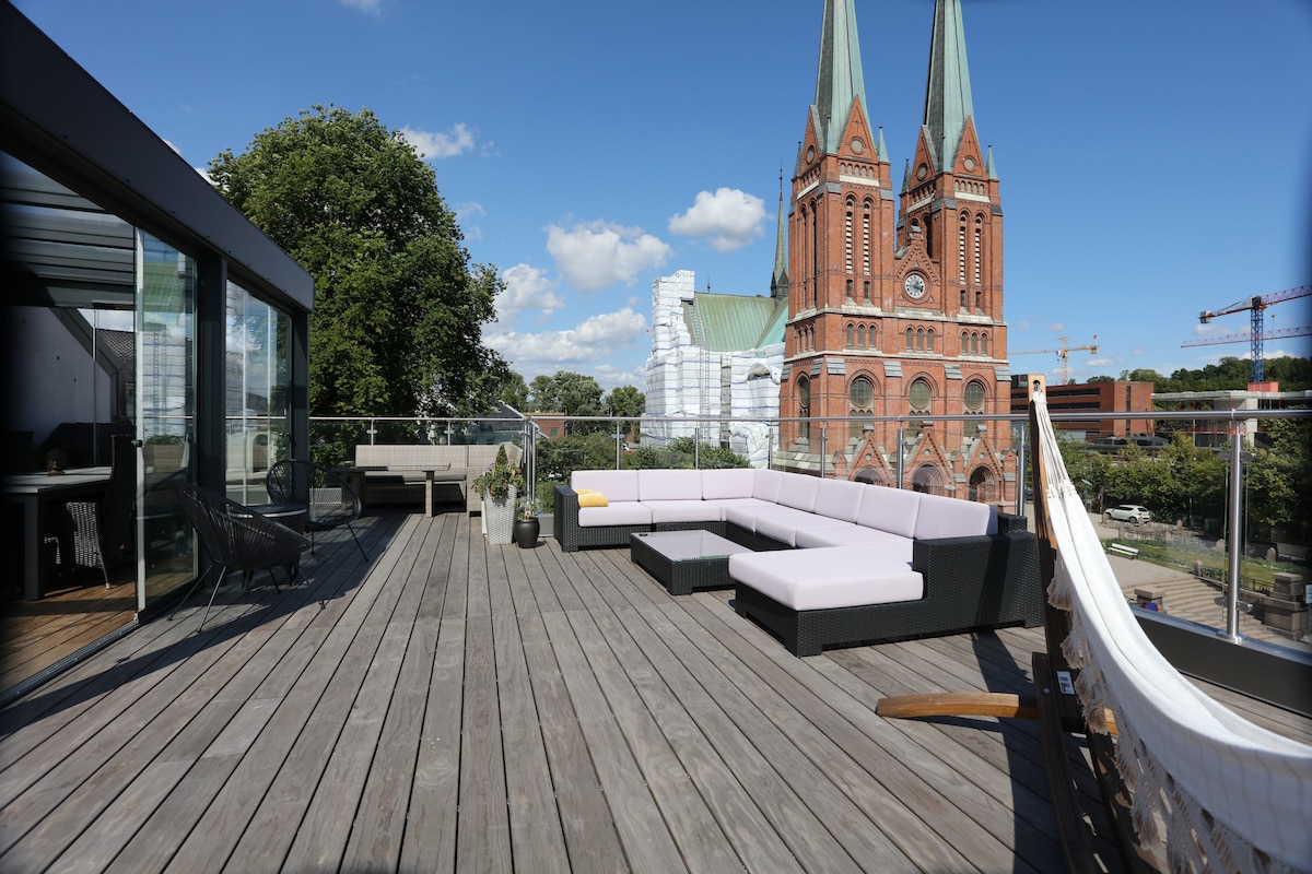 A spacious terrace features a sectional seating arrangement, complemented by a modern railing. The view includes a historic church with tall spires in the background, while greenery from nearby trees adds a touch of nature. The surface is made of wood, enhancing a contemporary outdoor setting.