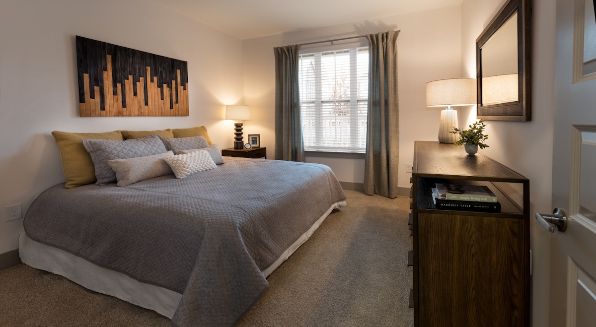 A serene bedroom features a spacious bed dressed in neutral tones, flanked by bedside tables with lamps. Light enters through a window dressed with sheer curtains, while a piece of wall art adds interest above the headboard. A dresser is positioned along one wall.