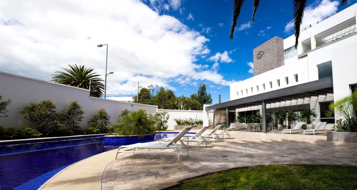 A modern outdoor space features a long, blue swimming pool surrounded by lounge chairs. Lush greenery and palm trees are visible along the perimeter. A shaded seating area is situated near the pool, with a white architectural structure in the background against a partly cloudy sky.