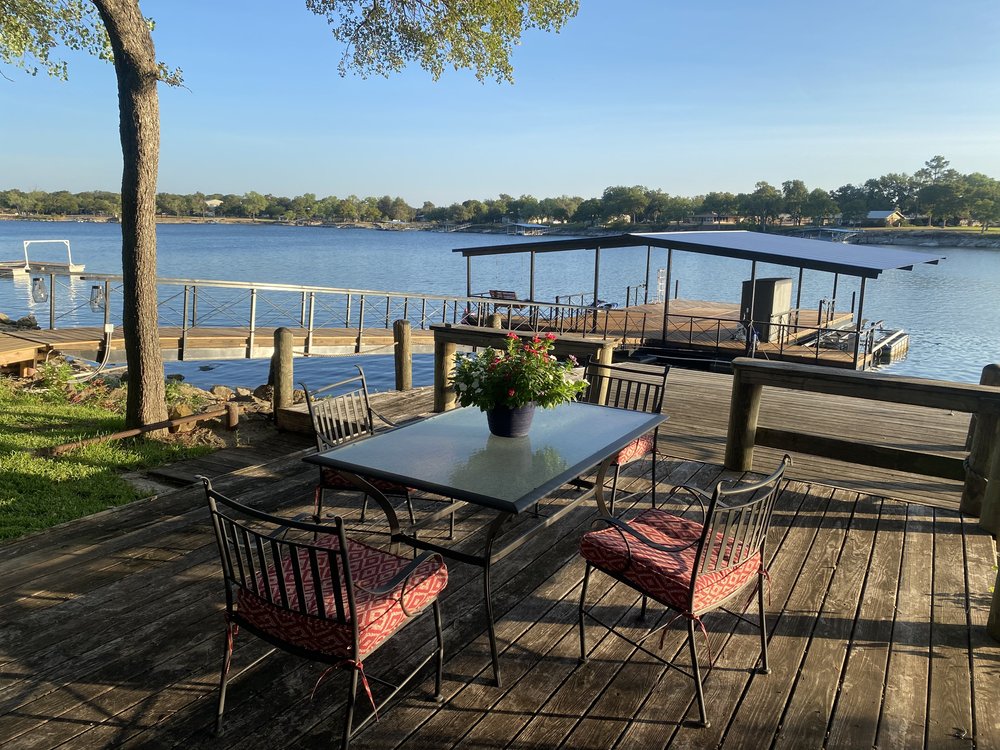 A waterfront patio is displayed, featuring a rectangular table surrounded by four chairs with cushioned seats. A potted flower arrangement sits on the table. In the background, a large dock extends into the serene lake, bordered by trees along the shore.