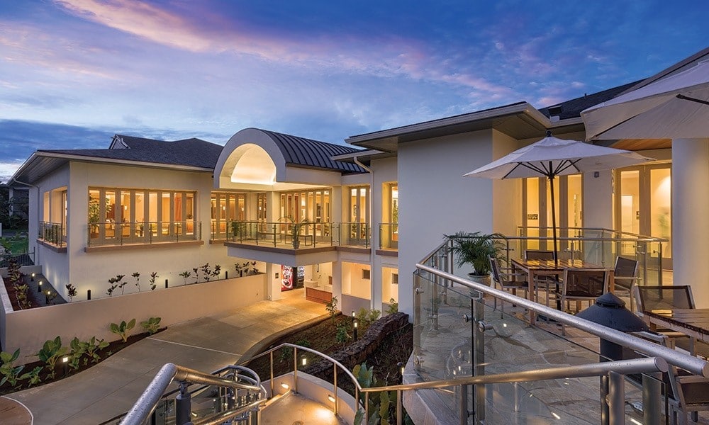 The image captures a modern resort building, designed with large glass windows that provide ample natural light. A spacious patio area is visible, featuring outdoor furniture and umbrellas. The structure is complemented by landscaped greenery, set against a tranquil twilight sky.
