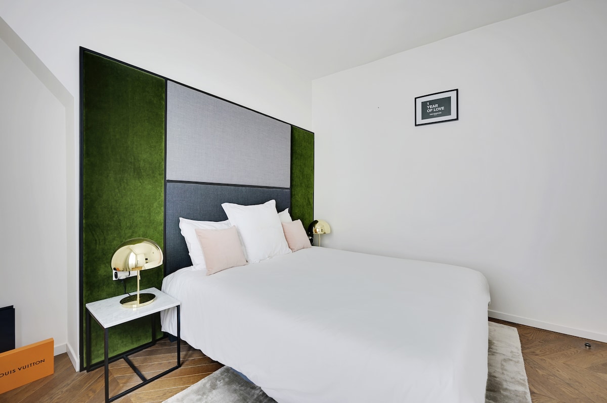 A spacious bedroom contains a comfortable queen-size bed with white linens. A stylish headboard in green and gray adds a modern touch. Two bedside lamps are positioned on either side of the bed, and a minimalist artwork is displayed on the wall above.