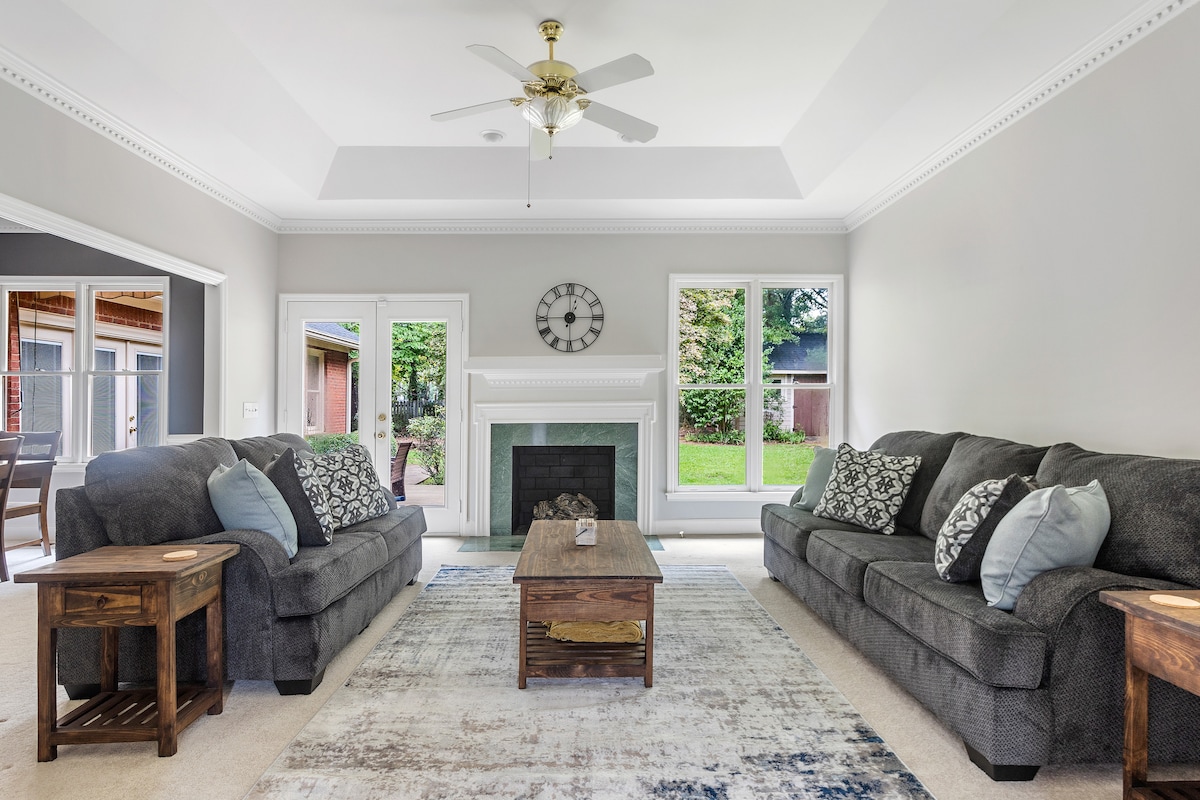 The living room features two spacious couches placed across from each other, with decorative cushions enhancing comfort. A wooden coffee table sits at the center, while large windows allow natural light to fill the space. A ceiling fan and a classic wall clock add functional touches.