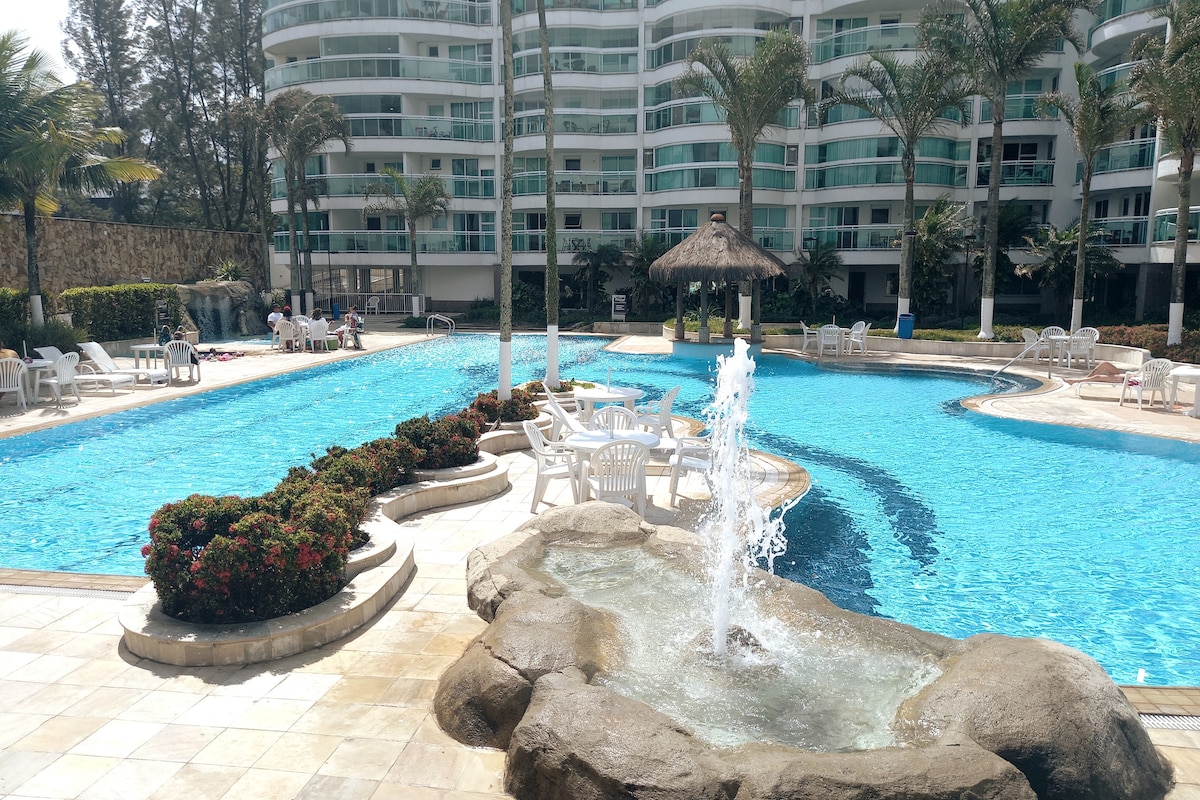 A large outdoor pool area is surrounded by well-maintained gardens and palm trees. A rock-styled fountain adds a natural touch, while several lounge chairs and umbrellas are positioned along the water's edge, inviting relaxation in a calm setting.