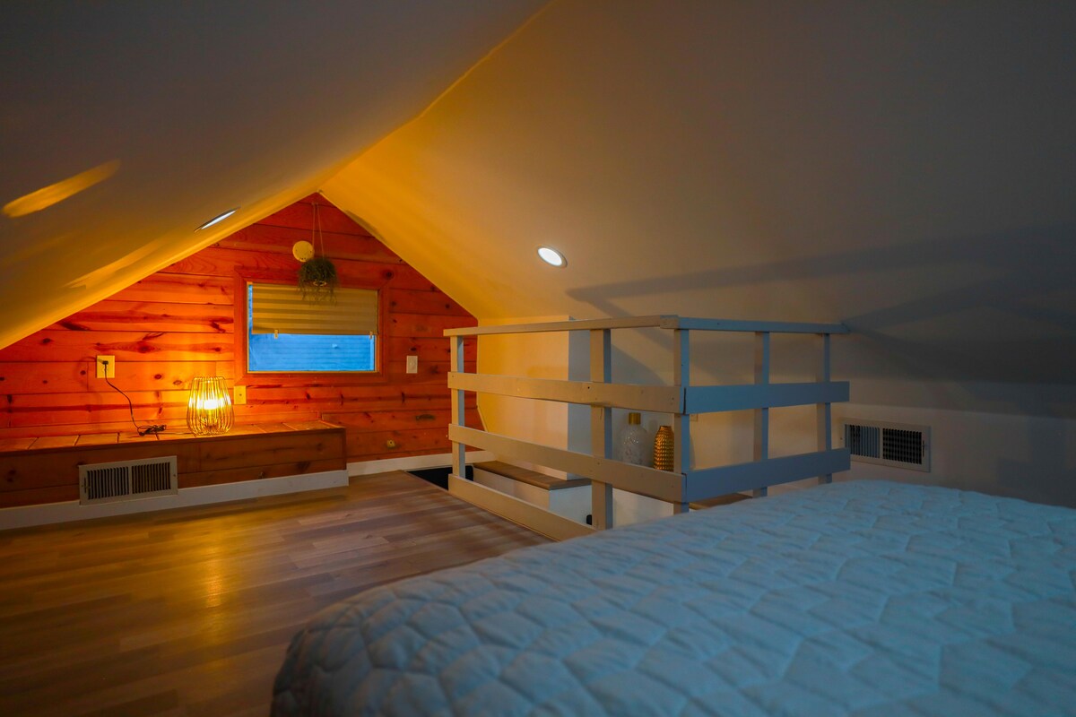 The loft area features wooden paneling against a softly lit backdrop, creating a warm ambiance. A cozy bed is positioned beneath a sloped ceiling, and a nearby window allows natural light to illuminate the space. Decorative elements, including light fixtures, add charm to the room.