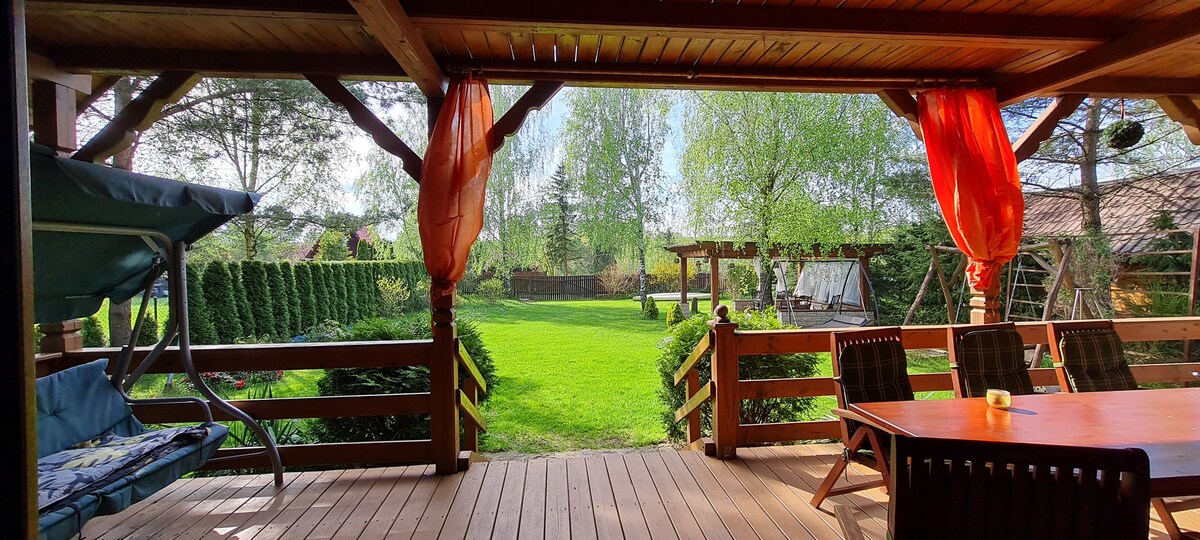 A wooden terrace overlooking a lush garden is framed by vibrant orange curtains. The space features a dining table and chairs, a swing seat, and an inviting view of the well-maintained green lawn, surrounded by trees and garden features.