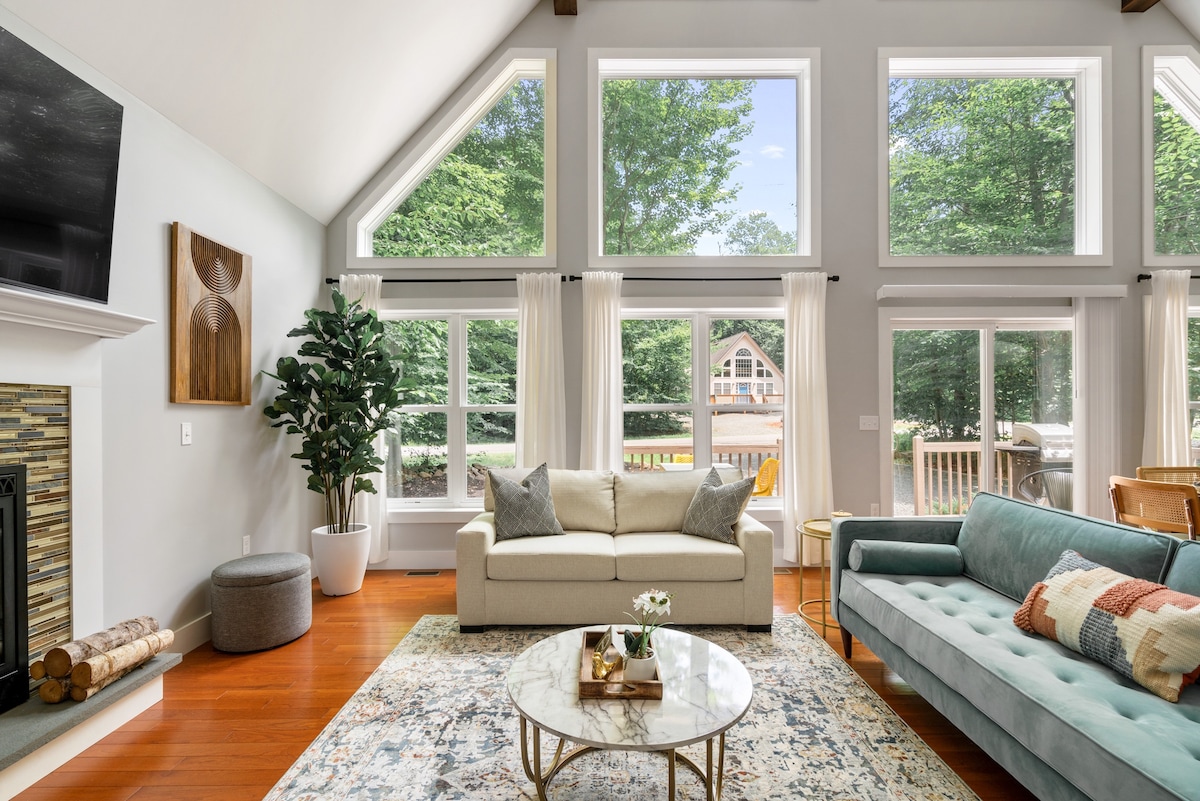 The living room features a high, vaulted ceiling with large windows allowing natural light to fill the space. A neutral sofa and a blue sofa are arranged around a round coffee table on a patterned rug. A gas fireplace and a decorative plant accent the cozy atmosphere.