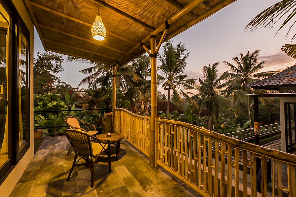 A covered balcony is shown featuring two woven chairs and a small table. Lush tropical plants and palm trees surround the area, with soft lighting illuminating the space. The horizon hints at a sunset, creating a serene outdoor setting.