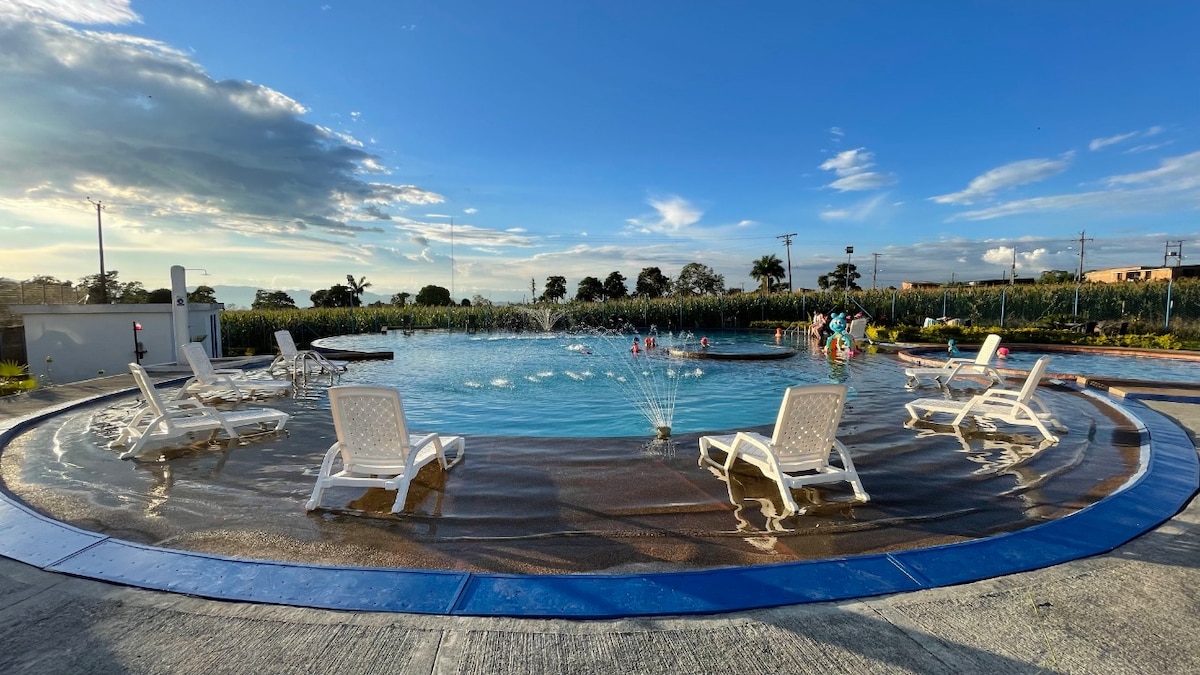 A spacious outdoor pool area is surrounded by lounge chairs, offering a relaxing environment. Water fountains feature playful jets in the center, while lush greenery and distant palm trees create a tranquil backdrop under a bright blue sky.