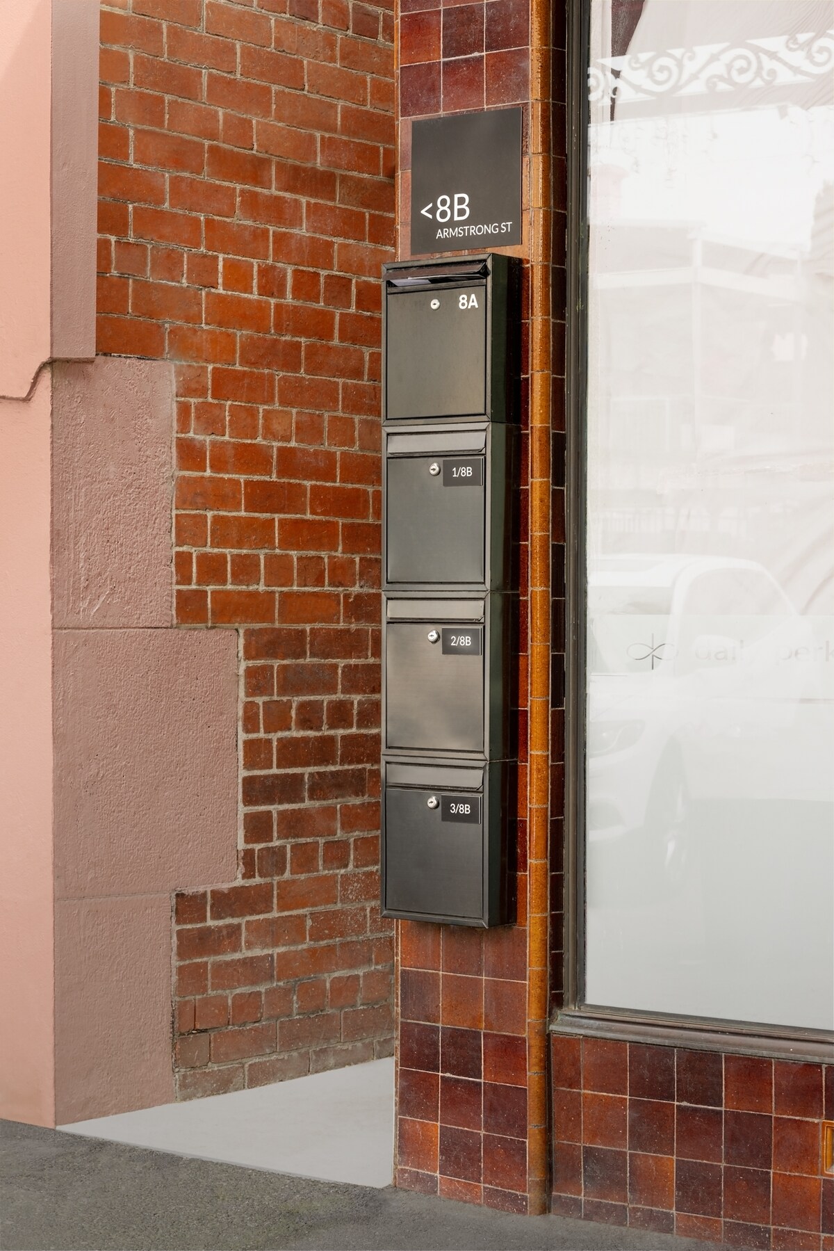 A brick wall features a series of four sleek black mailboxes, labeled with numbers. The design is modern, emphasizing functionality. The surrounding area includes a glimpse of a glass window, creating a blend of urban architecture.