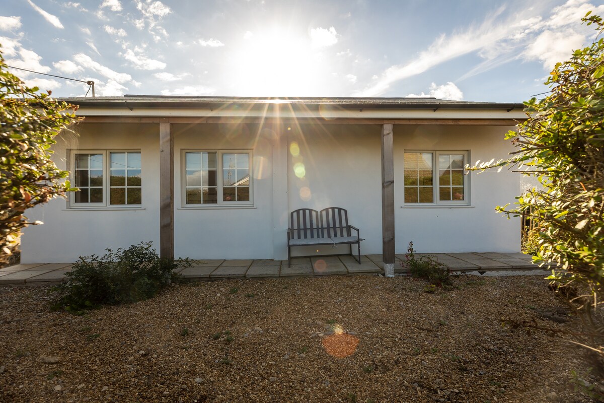 The cottage exterior is highlighted by soft sunlight streaming through a clear sky. A simple wooden bench is positioned on a stone patio, flanked by two large windows that illuminate the inviting interior.
