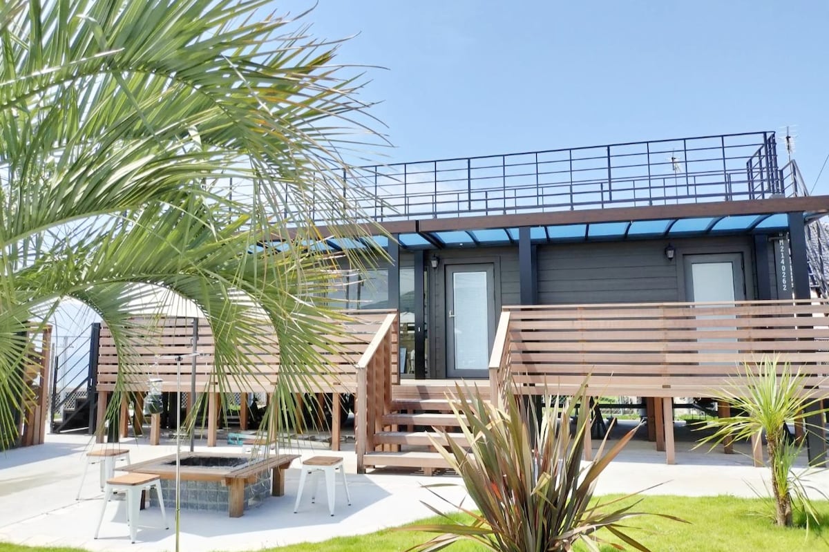 The vacation rental features a modern container house elevated on a raised platform, surrounded by tropical greenery. A wooden deck with a staircase leads to entry doors, while a spacious outdoor area is designed for relaxation and leisure. Large windows provide natural light.
