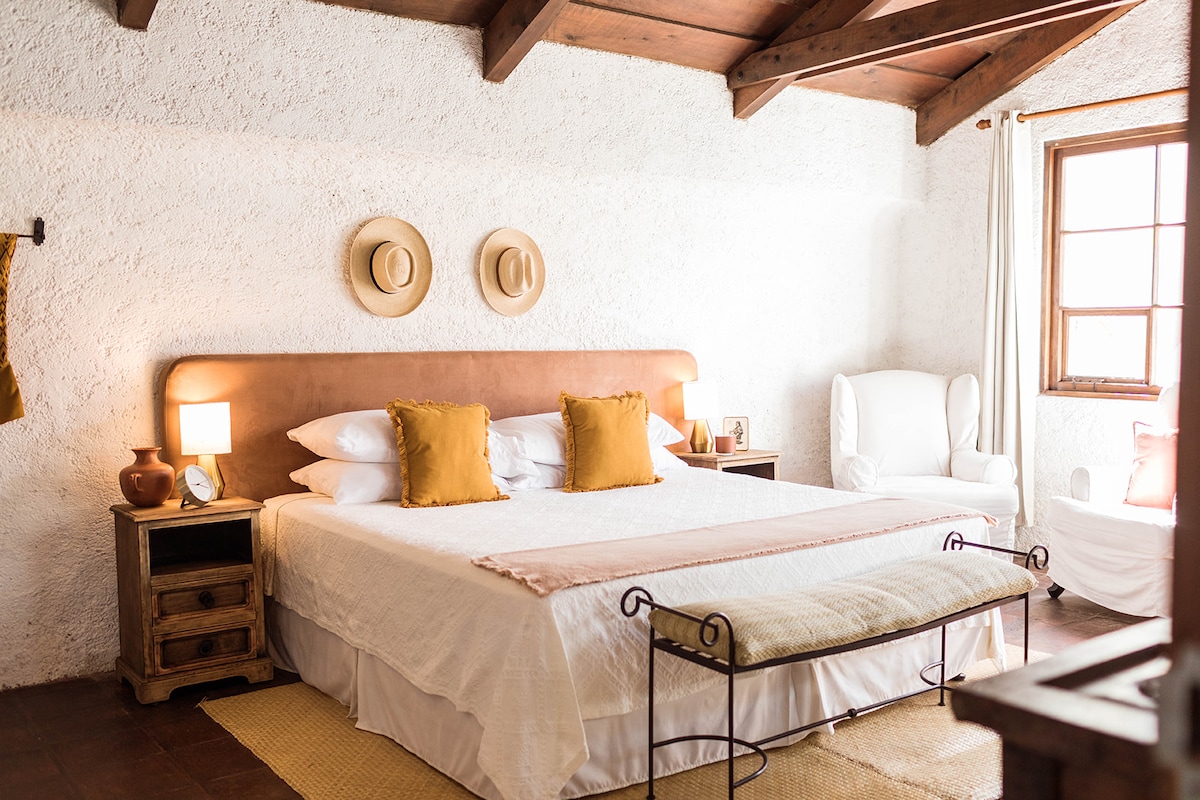 A spacious room is featured with a king size bed adorned with quality cotton sheets. Soft yellow pillows provide a welcoming touch. Natural light enters through a window, and two straw hats are hung on the wall. A cozy armchair is positioned nearby.