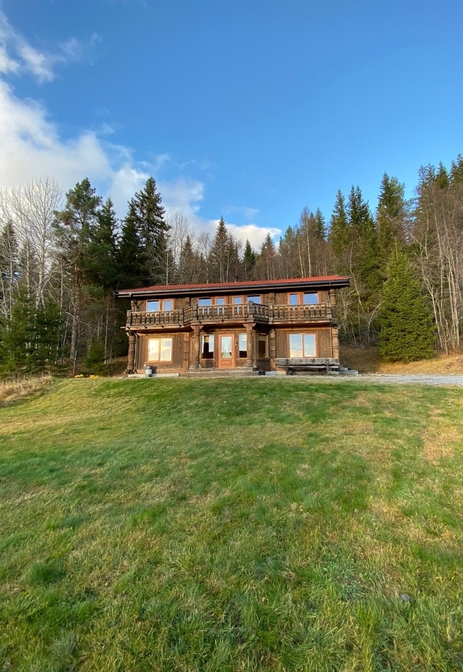 The two-story wooden cabin is set against a backdrop of trees, featuring large glass doors that provide an inviting entrance. The well-kept lawn surrounds the cabin, while a clear sky with scattered clouds is visible above.
