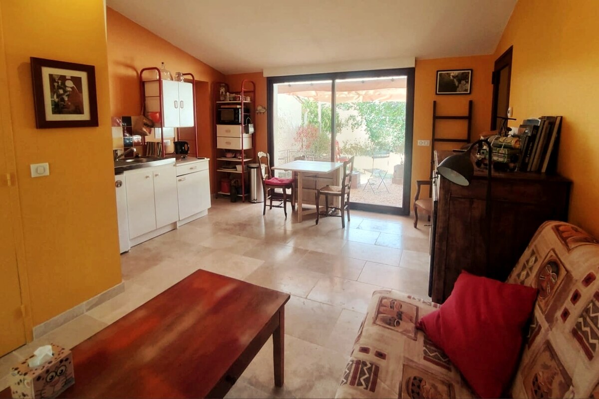 A bright studio space features warm orange walls and natural stone flooring. The kitchenette is equipped with essential appliances. A dining table set for four is positioned near large glass doors, allowing views of an outdoor area adorned with greenery. A cozy sitting area includes a convertible sofa.