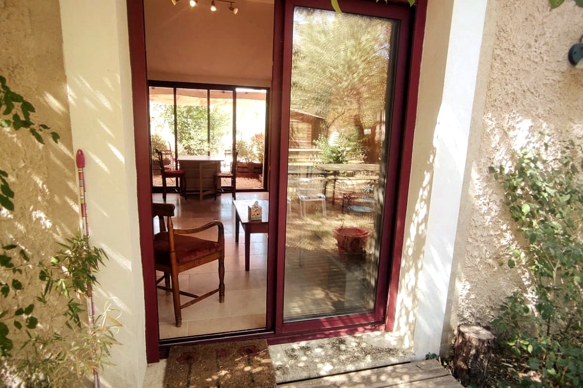 A double glass door provides access to the studio, framed by greenery. The door opens to a spacious area illuminated by natural light, revealing a blend of indoor and outdoor elements, with a view of a patio area beyond.