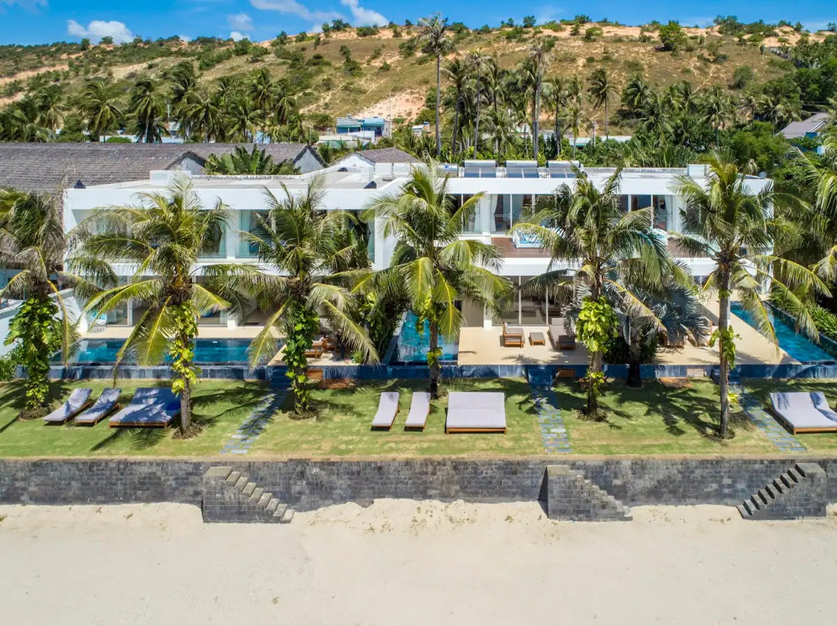 A modern villa is positioned near the beach, surrounded by palm trees. The spacious lawn features lounge chairs and pathways leading to a private pool. The architecture showcases large glass windows, emphasizing a seamless connection with the outdoor landscape.