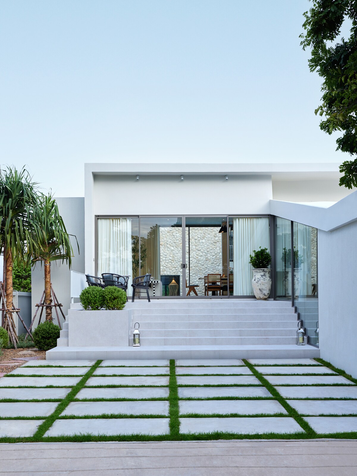 A modern exterior features a well-defined entrance with steps leading up to large glass sliding doors. Lush greenery lines the sides, complemented by potted plants. The clean lines of the concrete pathway are accented by neatly arranged grass, creating a serene outdoor atmosphere.