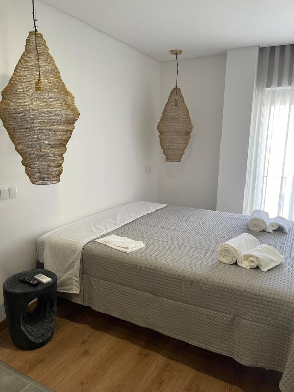 A well-lit sleeping area features a spacious bed covered with a light grey blanket. Two woven pendant lights hang from the ceiling, and neatly rolled towels are placed on the bed. A small, dark stool is positioned beside the bed, complementing the wooden floor.