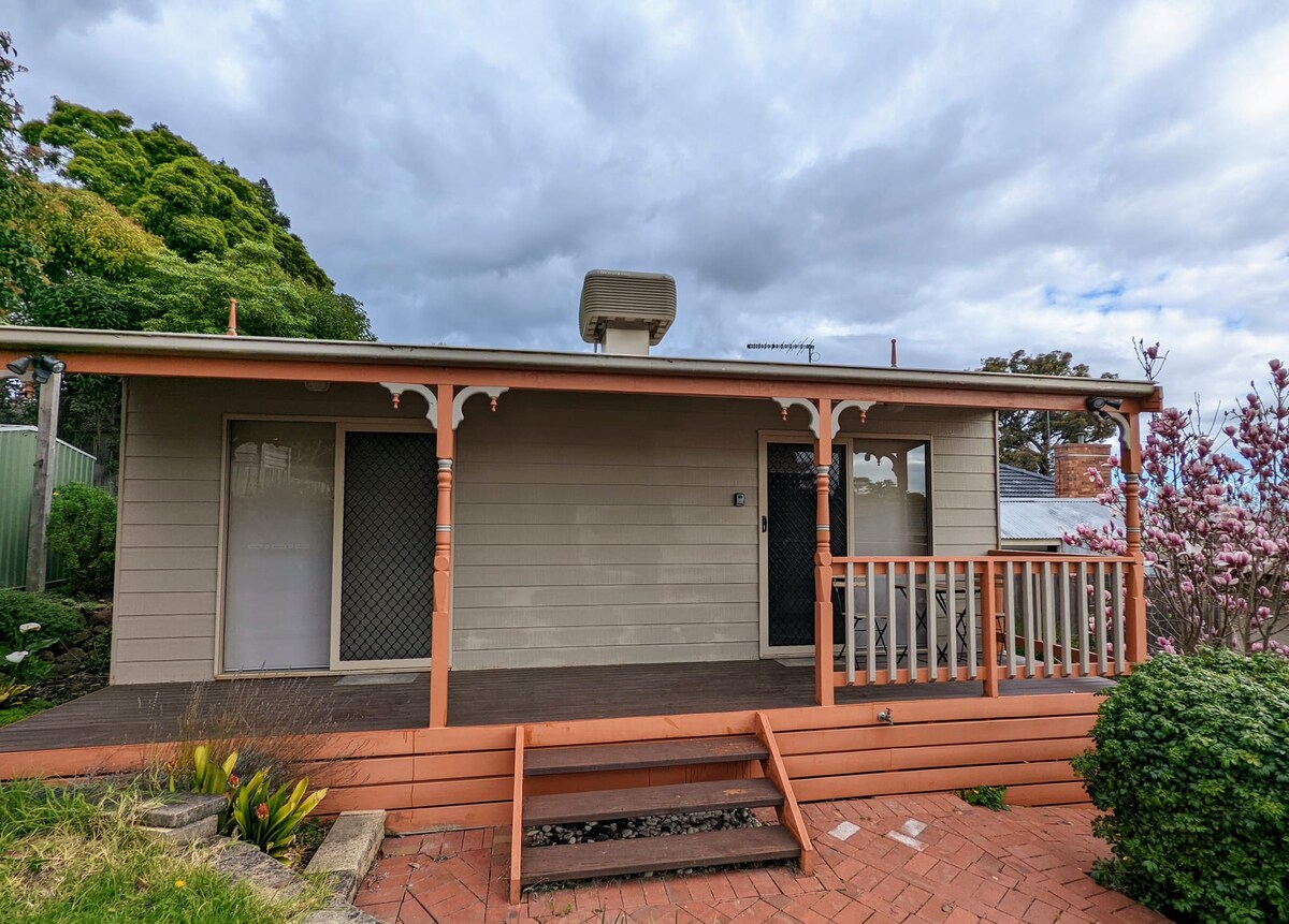 The exterior of a one-bedroom cottage is presented, featuring a welcoming porch with steps leading up. The facade is adorned with decorative trim, while large windows on either side offer views into the interiors. Lush greenery and blooming plants are visible in front of the cottage.