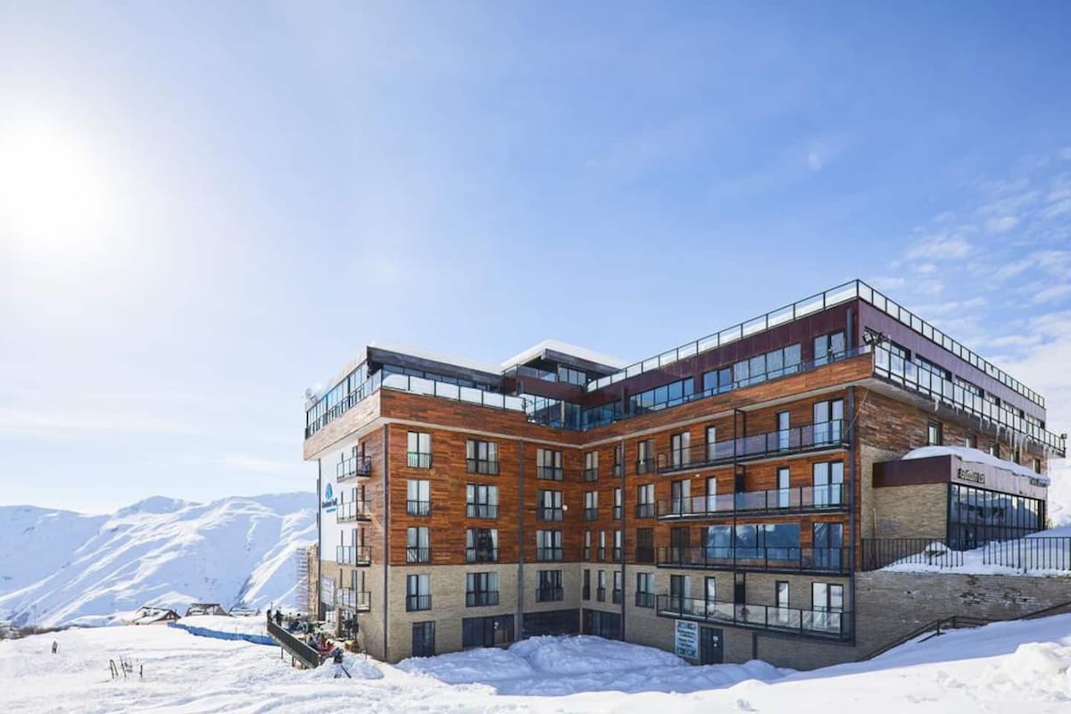 The modern hotel structure features multiple levels with large windows and wooden accents, blending into the surrounding snowy landscape. Sunlight reflects off the snow, creating a bright and inviting ambiance. Balconies are visible on several floors, offering scenic views of the mountains.