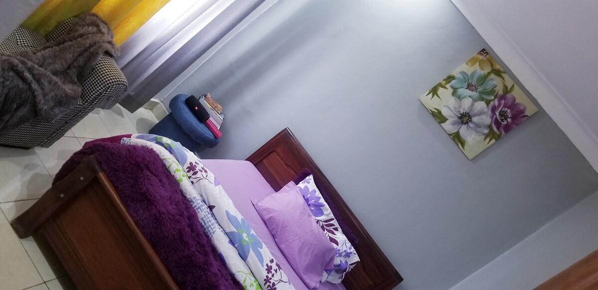 A cozy bedroom is presented with a wooden bed featuring floral bedding in soft pastels. A plush purple throw is draped across the bed. A patterned chair sits in the corner, and a vibrant floral artwork hangs on the wall, complemented by light grey and yellow curtains.