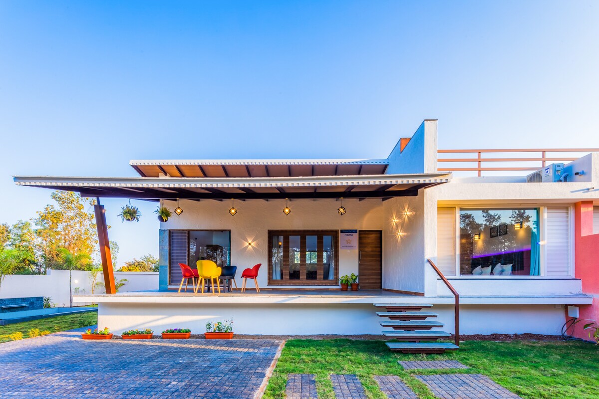 The villa's exterior features a modern design with a welcoming porch, furnished with colorful chairs and a table. The landscaped green area complements the inviting entrance. Large windows allow for natural light to fill the interior spaces, while the wide steps lead to the main entrance.