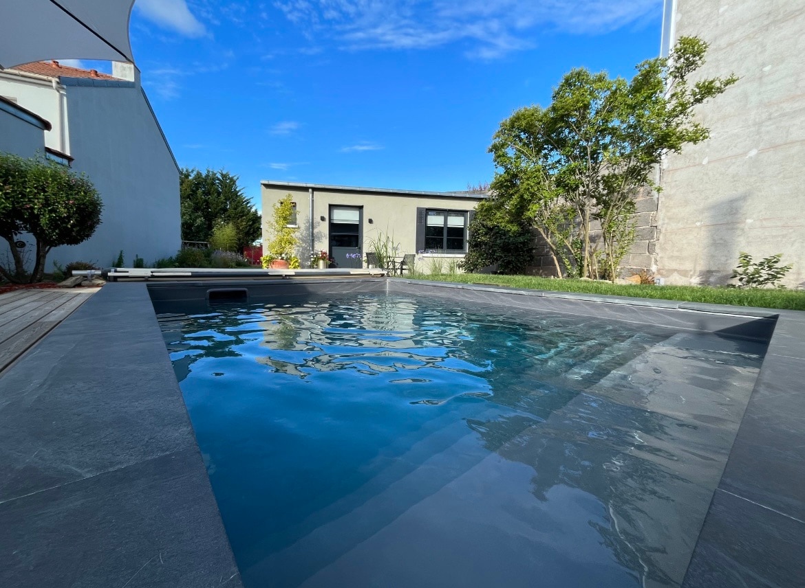 A tranquil swimming pool is set within a sleek stone surface, reflecting the blue sky. Surrounding greenery provides a natural backdrop, while the nearby building and garden area offer a serene setting for relaxation.