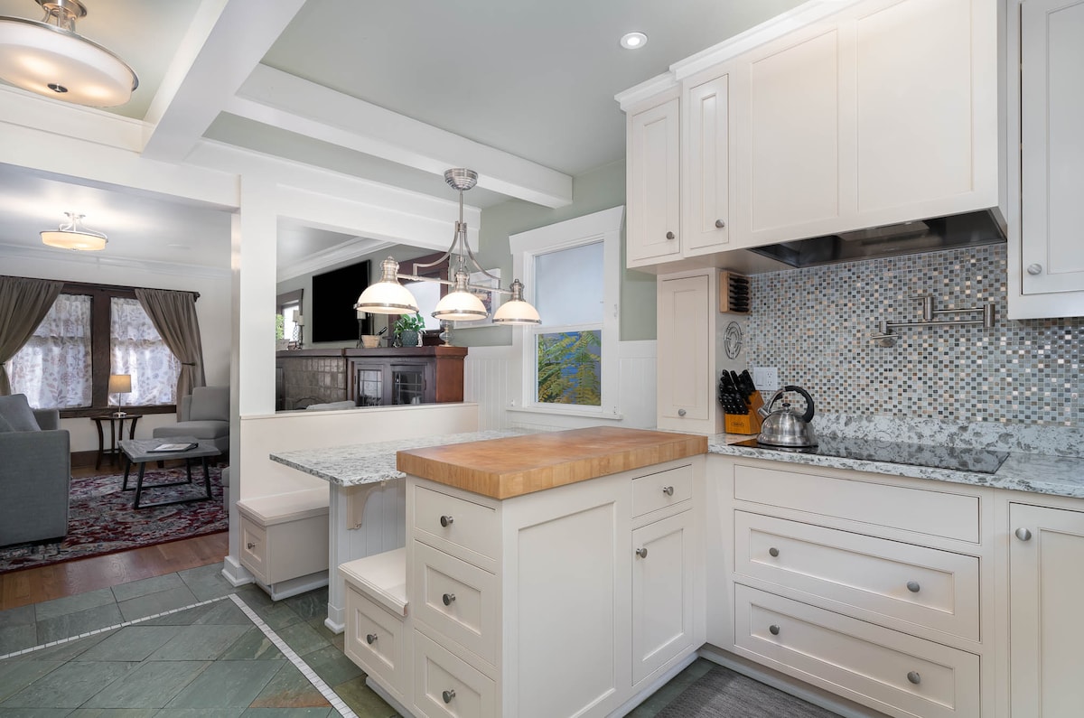 The kitchen features a modern design with light cabinetry and a central butcher block island. Textured tile backsplash complements the granite countertops. Pendant lights illuminate the workspace, while a dining area with a table for four is visible through the adjacent archway.