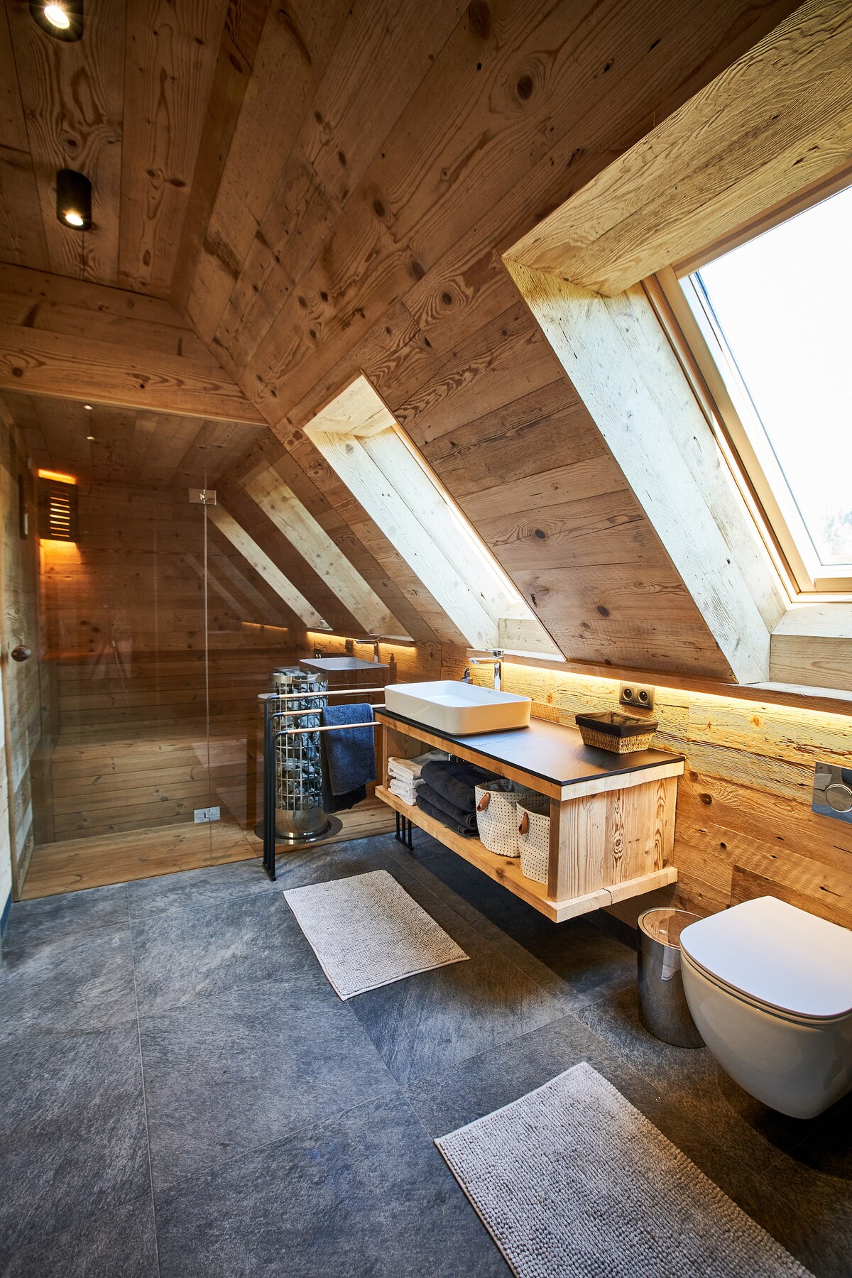 A modern bathroom is presented with sloped wooden walls and skylight windows allowing natural light to enter. A spacious wooden vanity is accompanied by a washbasin, with open shelving displaying neatly arranged towels. A glass shower enclosure complements the clean design, with minimalistic floor mat placement.