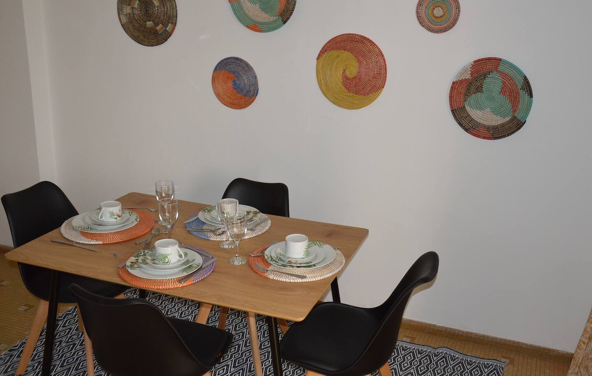 A dining area features a wooden table set for four, accompanied by black chairs. Decorative wall art made of woven materials adds color to the white wall. Glassware and dishware are arranged on colorful placemats, creating a welcoming atmosphere for dining.
