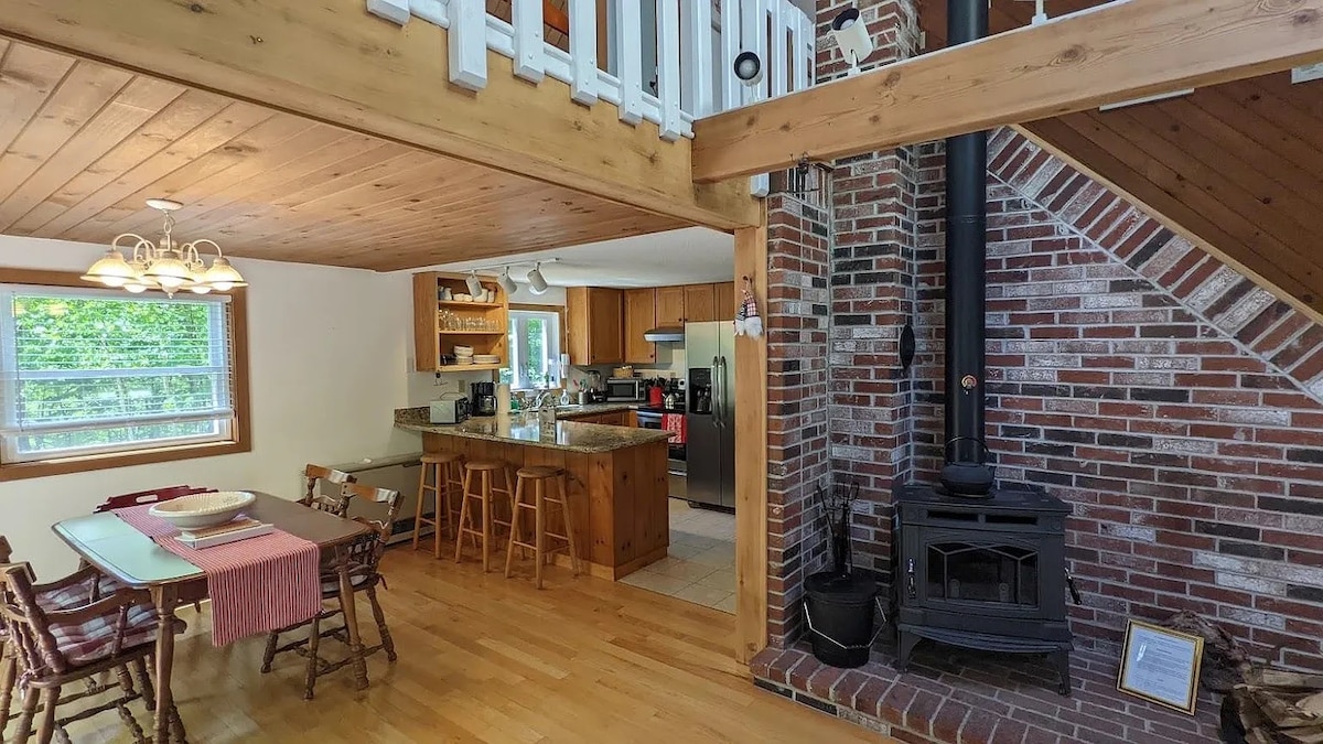 An open-plan kitchen and dining area feature wooden floors and a central island with bar seating. A wood-burning stove is positioned against a brick wall, adding warmth to the space. Natural light enters through multiple windows, enhancing the inviting atmosphere.