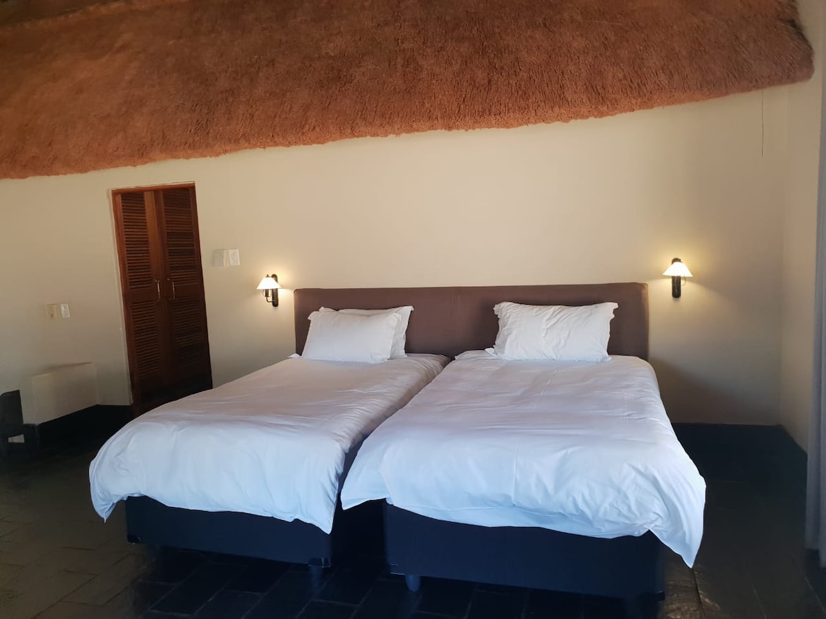 A bedroom features two spacious beds covered with white linens, positioned against a neutral wall. Soft lighting is provided by wall sconces on either side. A wooden closet is visible in the background, and the ceiling showcases a textured, natural material.