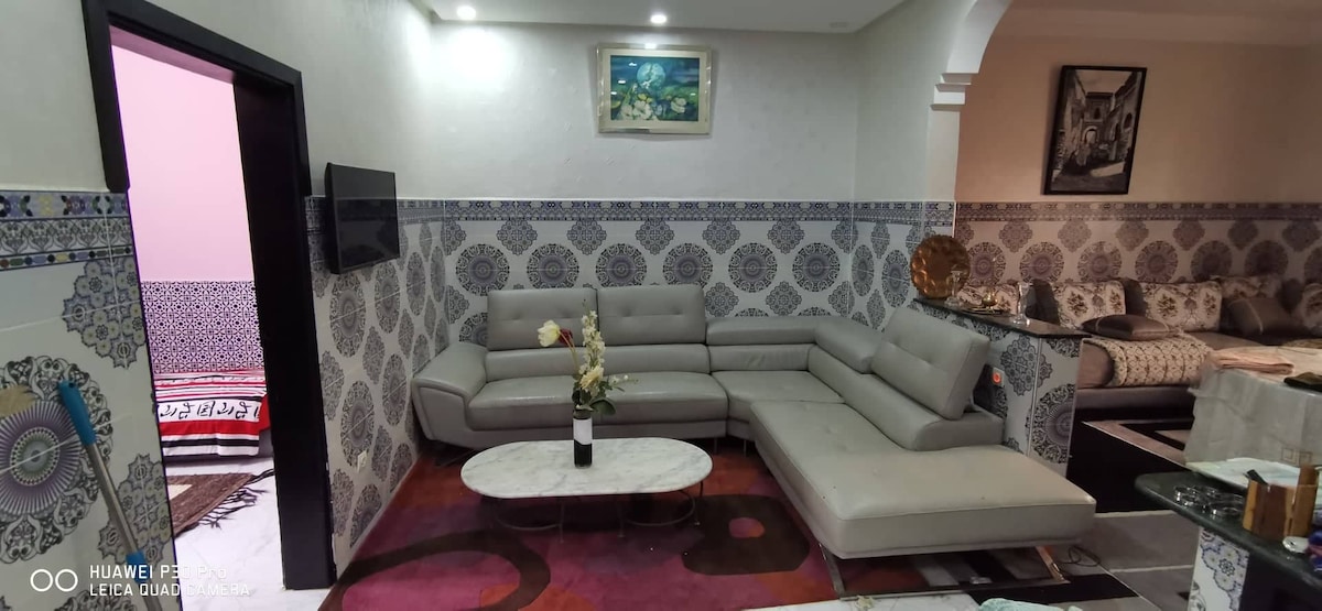 A modern living area is featured, showcasing a spacious sectional sofa in light grey. A circular marble coffee table sits in the center, while decorative floral arrangements add subtle accents. The walls are adorned with intricate tile patterns, and a flat-screen TV is mounted for entertainment.