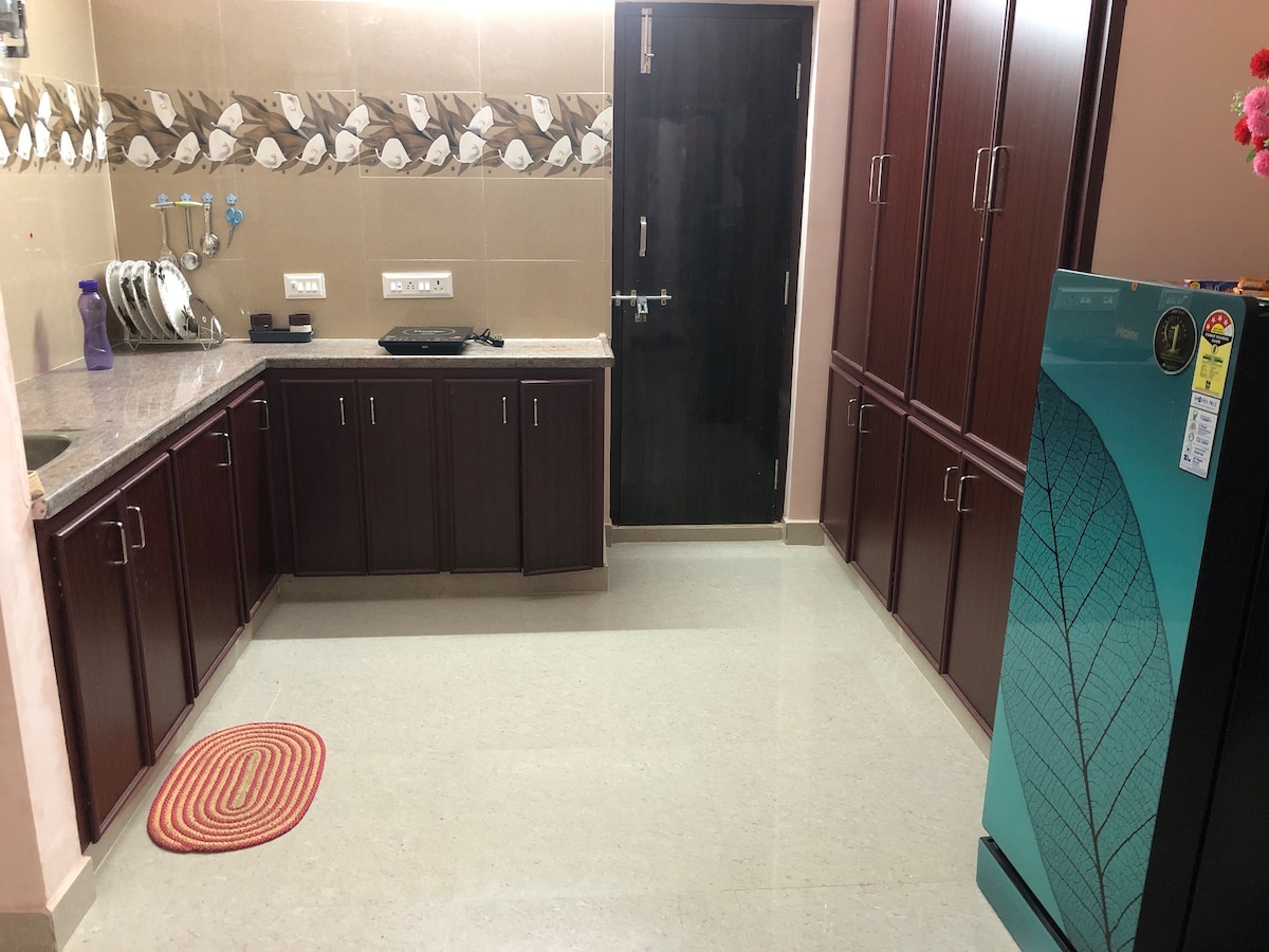 A modern kitchen features wooden cabinetry with ample storage space. The countertop is composed of light-colored stone, complemented by a backsplash with a subtle design. A refrigerator in teal is visible, alongside essential kitchen utensils and a round rug placed on the floor.