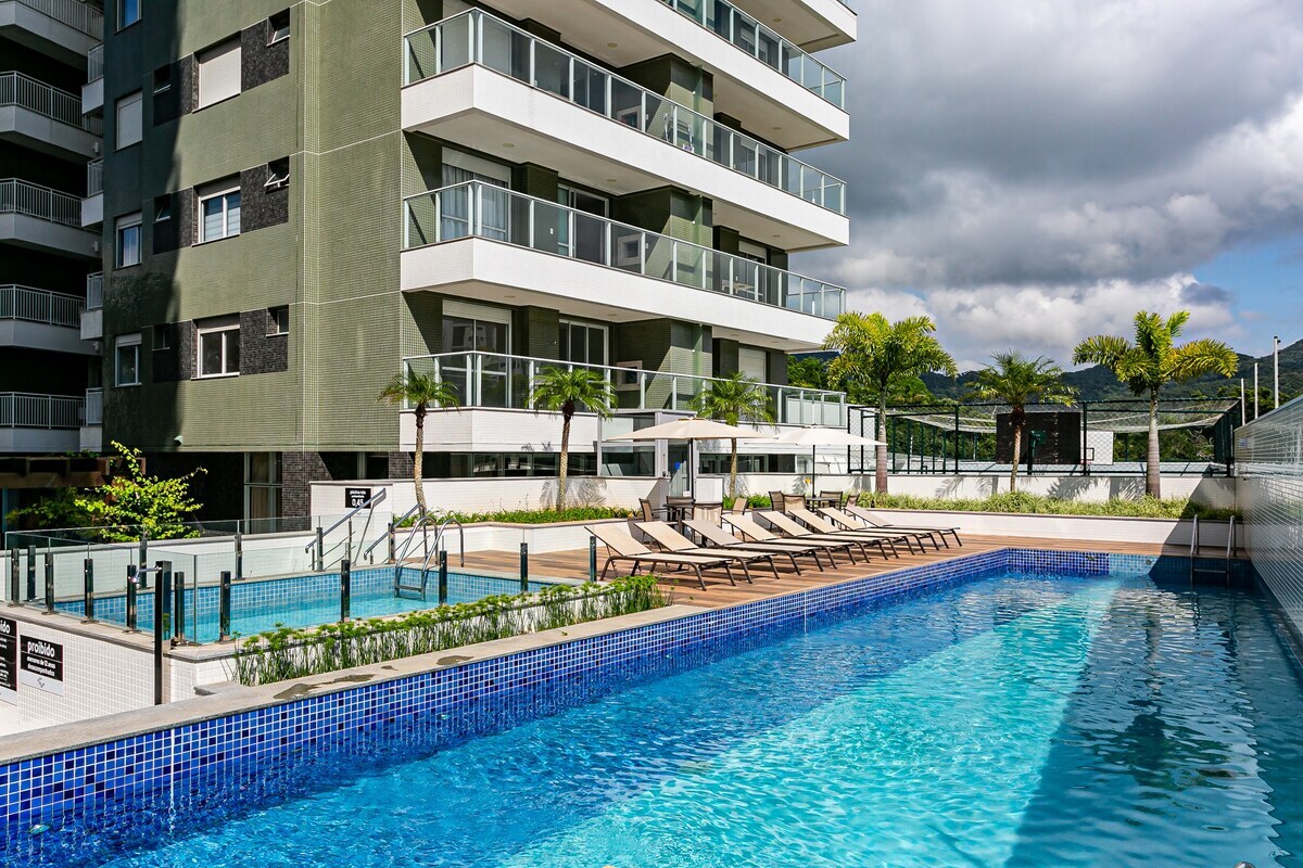 A well-maintained outdoor pool area is featured, surrounded by lounge chairs along the deck. Lush landscaping and modern architecture are visible, with multiple levels of the building creating a layered effect against a cloudy sky.