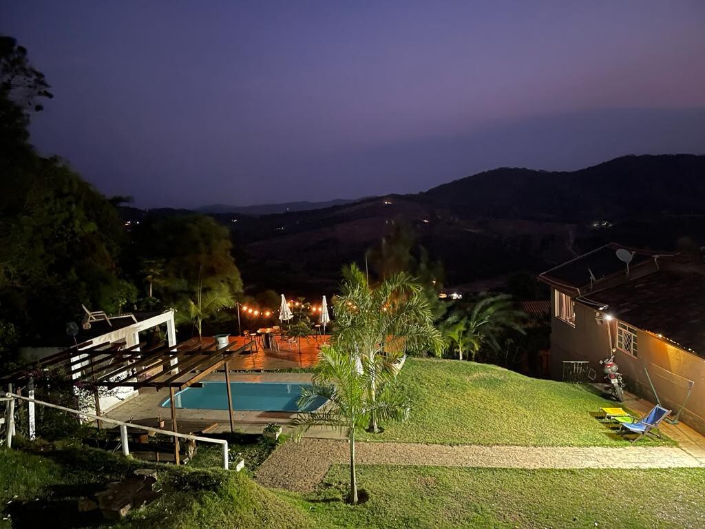 A serene nighttime view captures the pool area and deck, surrounded by lush greenery. The softly lit deck invites relaxation, while distant hills create a tranquil backdrop. The setting emphasizes a connection with nature, enhanced by ambient lighting.
