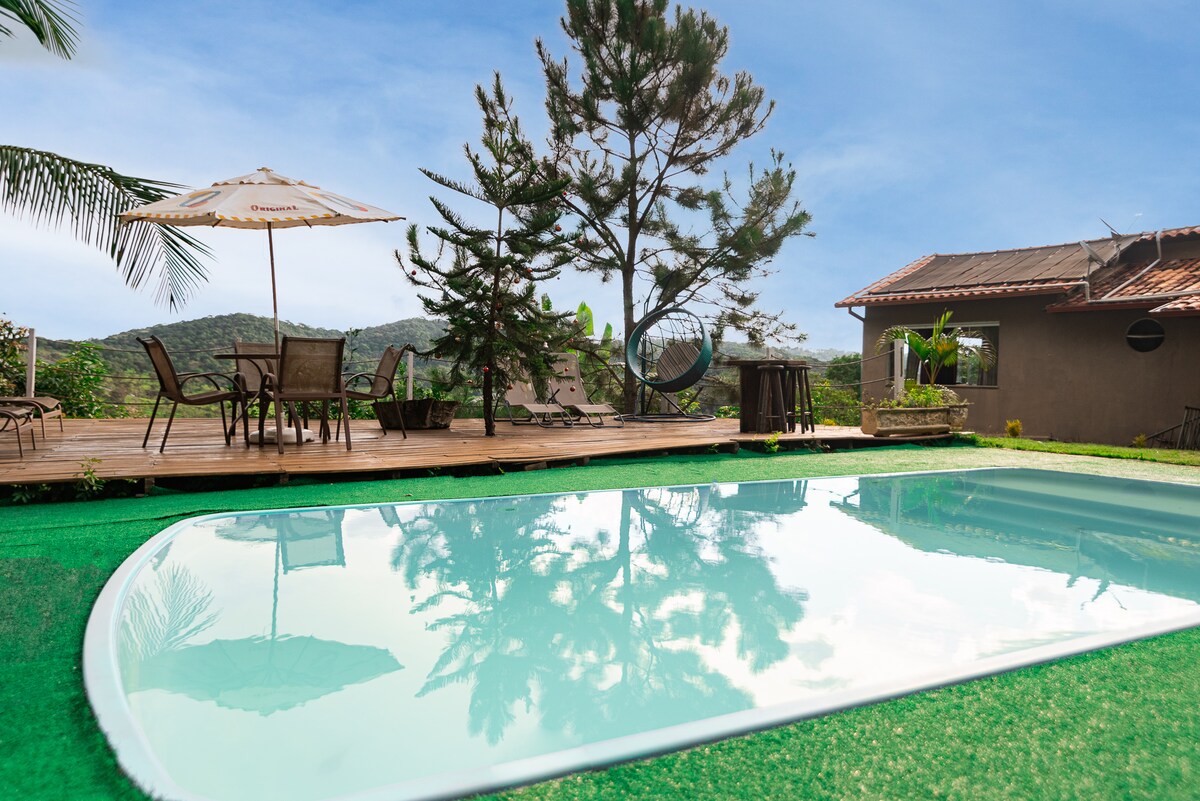 A serene outdoor area features a swimming pool surrounded by a lush green lawn. Wooden lounge chairs and a dining table with an umbrella can be seen on the deck, along with a vibrant tree and distant hills, creating a peaceful natural setting.
