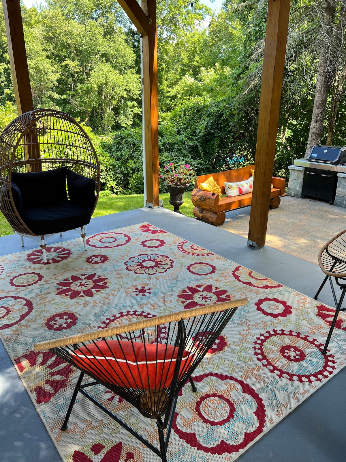 A covered patio area features plush seating arrangements on a colorful rug with floral patterns. A swinging chair adds a playful touch, while a stone grill stands ready for outdoor cooking against a backdrop of lush greenery.