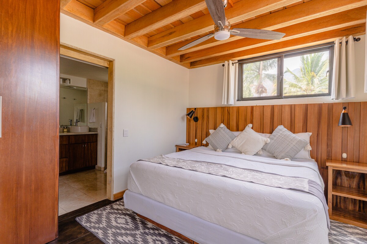 A spacious bedroom features a large bed with decorative pillows and a neutral-colored bedspread. Natural light enters through a large window, highlighting the wooden ceiling and floor. A doorway to the bathroom is visible, emphasizing an open layout.