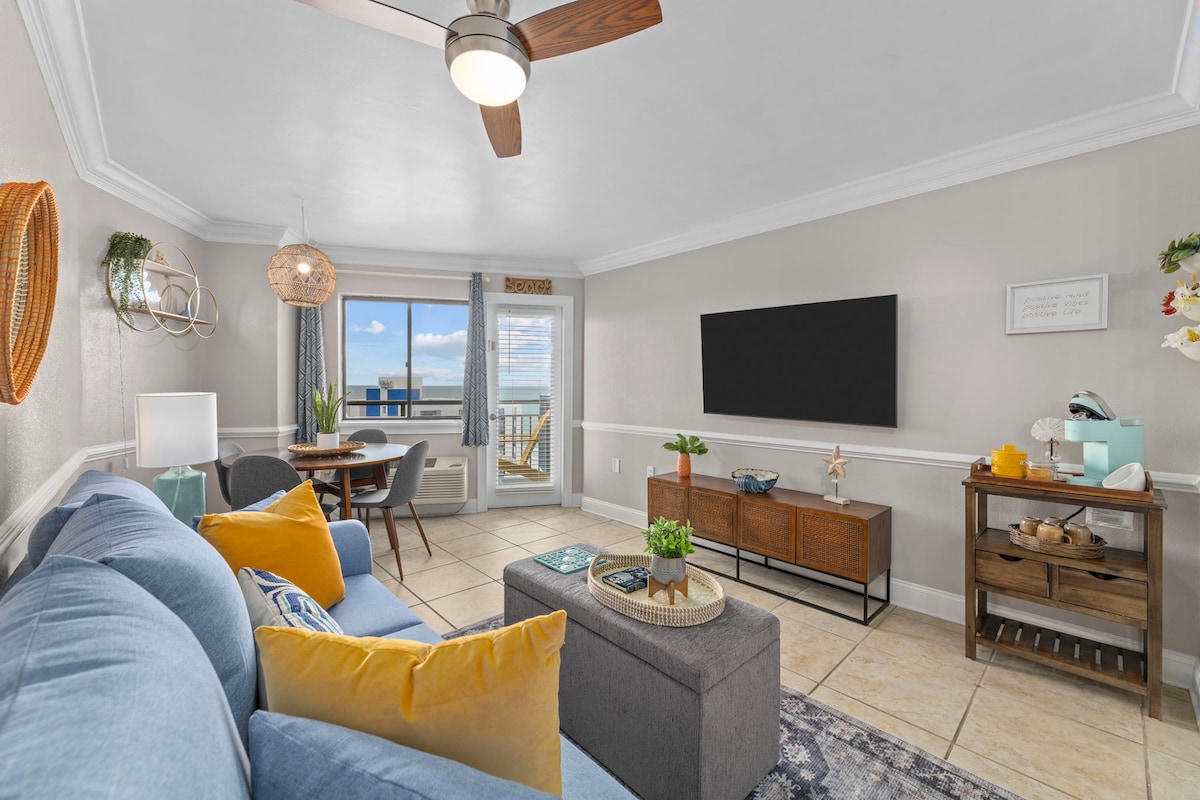 A cozy living area features a blue sofa adorned with bright yellow pillows. A coffee table sits in front, complemented by a wooden console and a potted plant. A large TV is mounted on the wall, while sliding glass doors provide a view of the ocean and lead to the private balcony.