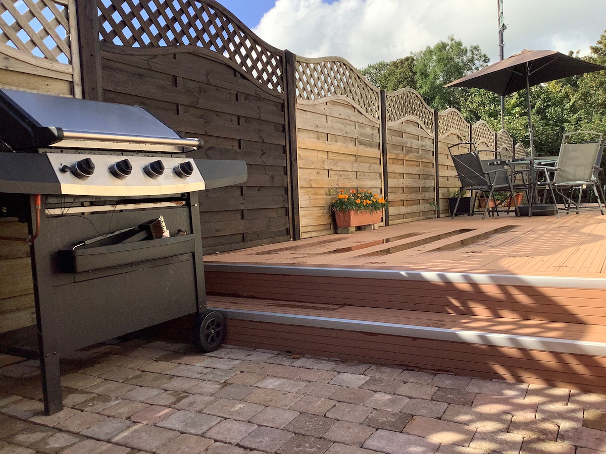 An outdoor deck area is shown, featuring a charcoal grill on wheels and a dining set covered by an umbrella. The space is enclosed by wooden lattice panels, while planters with flowers add a touch of greenery.