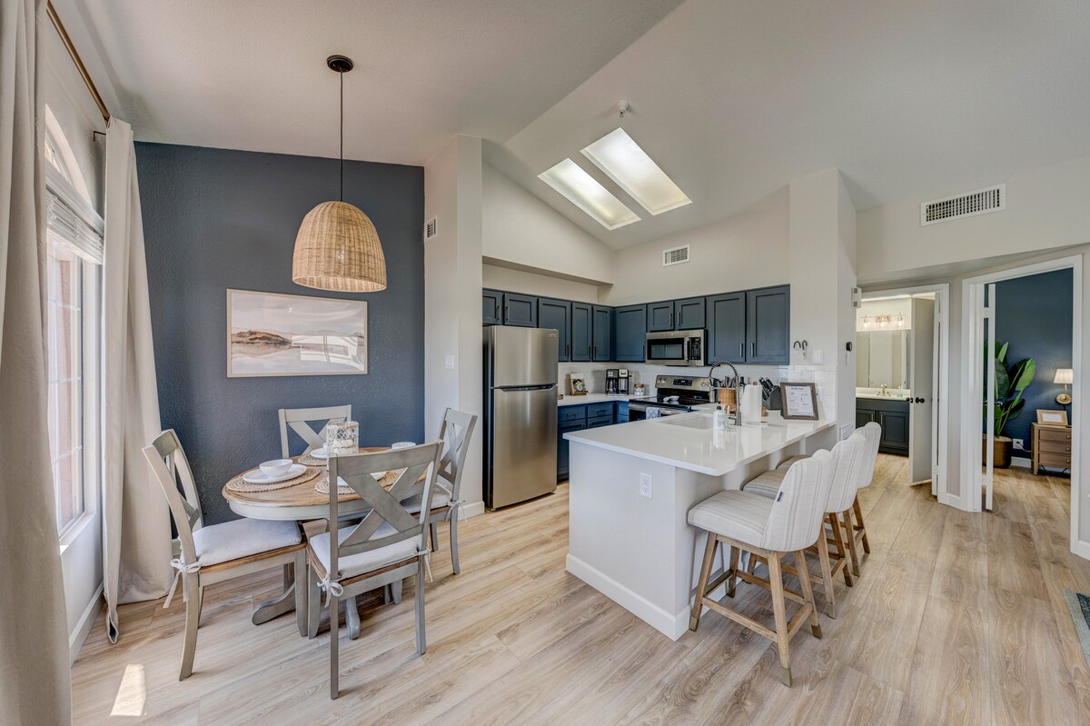 An open-concept kitchen and dining area is featured, showcasing light wood flooring and dark cabinetry. A round dining table is surrounded by four chairs, and the kitchen includes stainless steel appliances. Natural light enters through large windows, enhancing the inviting space.
