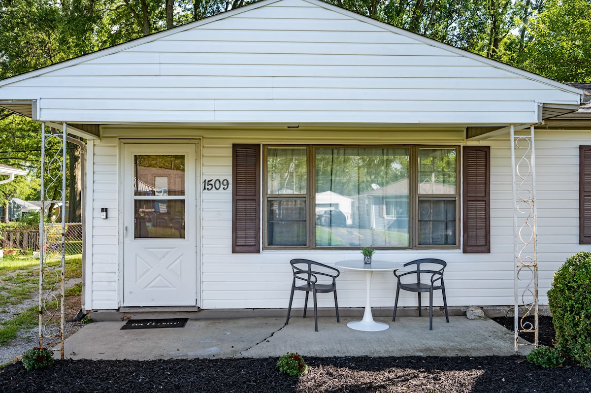 A welcoming entrance features a small outdoor seating area with two chairs positioned around a table. The light-colored facade is complemented by large windows adorned with brown shutters, offering a view into the home. A simple pathway leads to the front door.